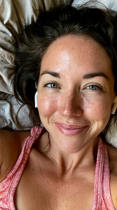 Freckled woman smiling in bed wearing earbuds