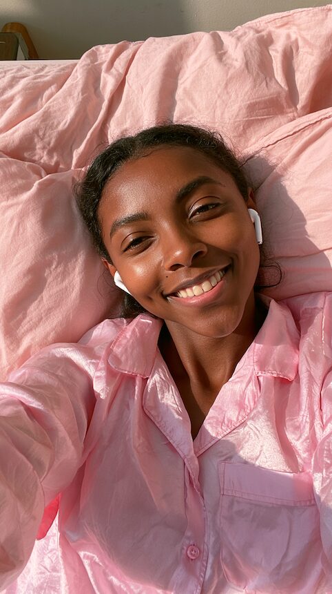 Smiling woman in pink pyjamas lying on pink bedding with earbuds