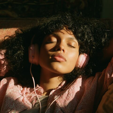 Woman resting with her eyes closed, listening to a NeuGlow session through pink headphones in warm evening light
